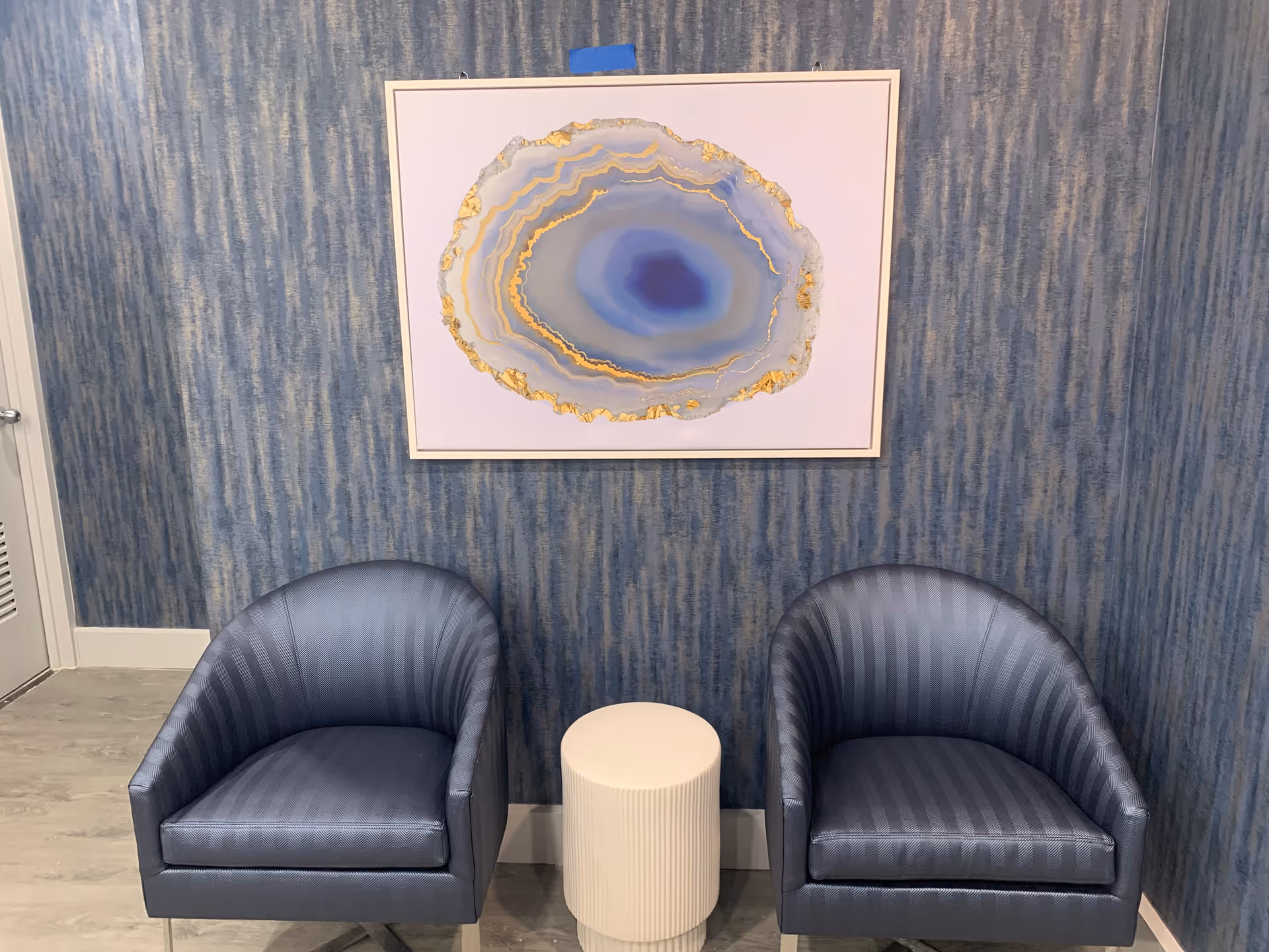 Two blue upholstered armchairs with a small white round table between them, set against a blue textured wall with a framed abstract artwork featuring blue and gold tones hanging above.