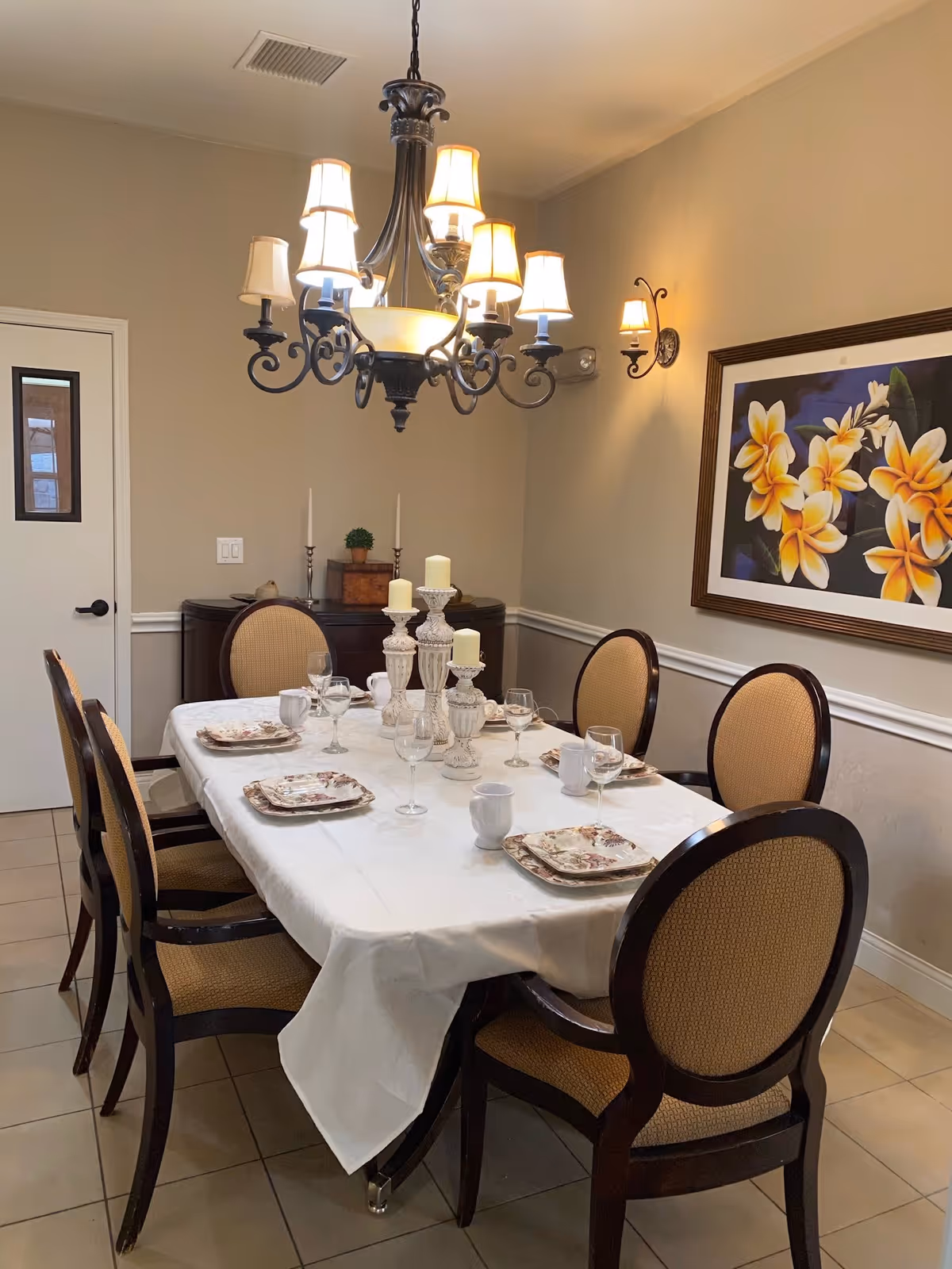 A dining room with a rectangular table covered with a white tablecloth, set with plates, cups, and wine glasses. Six upholstered chairs with wooden frames surround the table. A chandelier with multiple lampshades hangs above the table. On the wall, there is a framed picture of yellow and white flowers. A sideboard with decorative items is against the back wall.