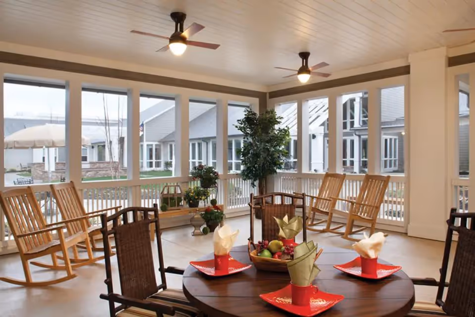 A bright and airy enclosed porch area with large windows overlooking an outdoor courtyard. The room features wooden rocking chairs along the walls, a round wooden table set with red plates and folded napkins, ceiling fans with lights, and potted plants adding greenery to the space.