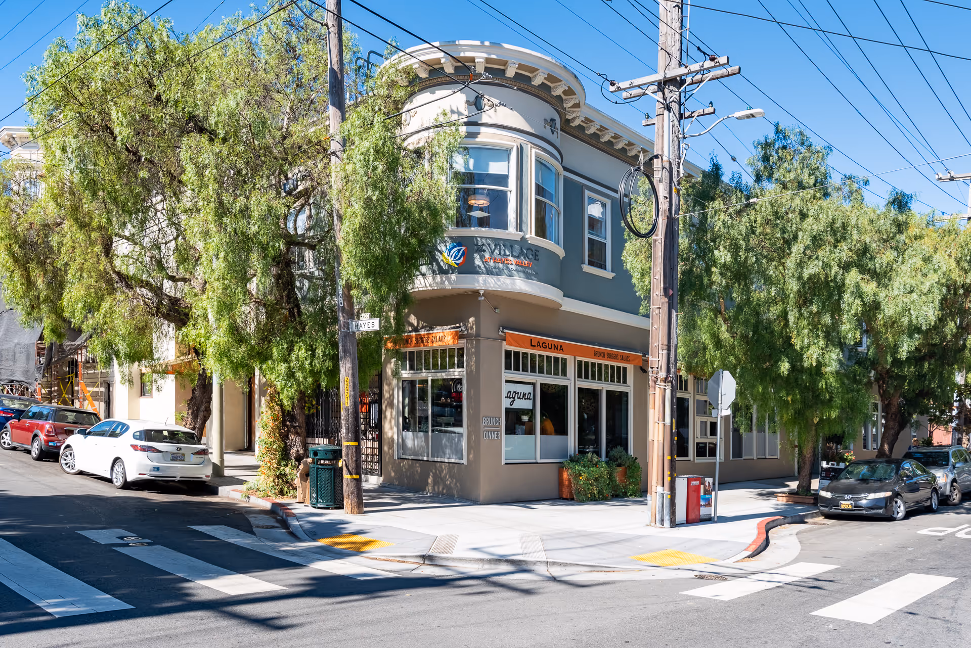 Street-level view of a two-story corner building with rounded bay windows, trees, and parked cars in front of a storefront.