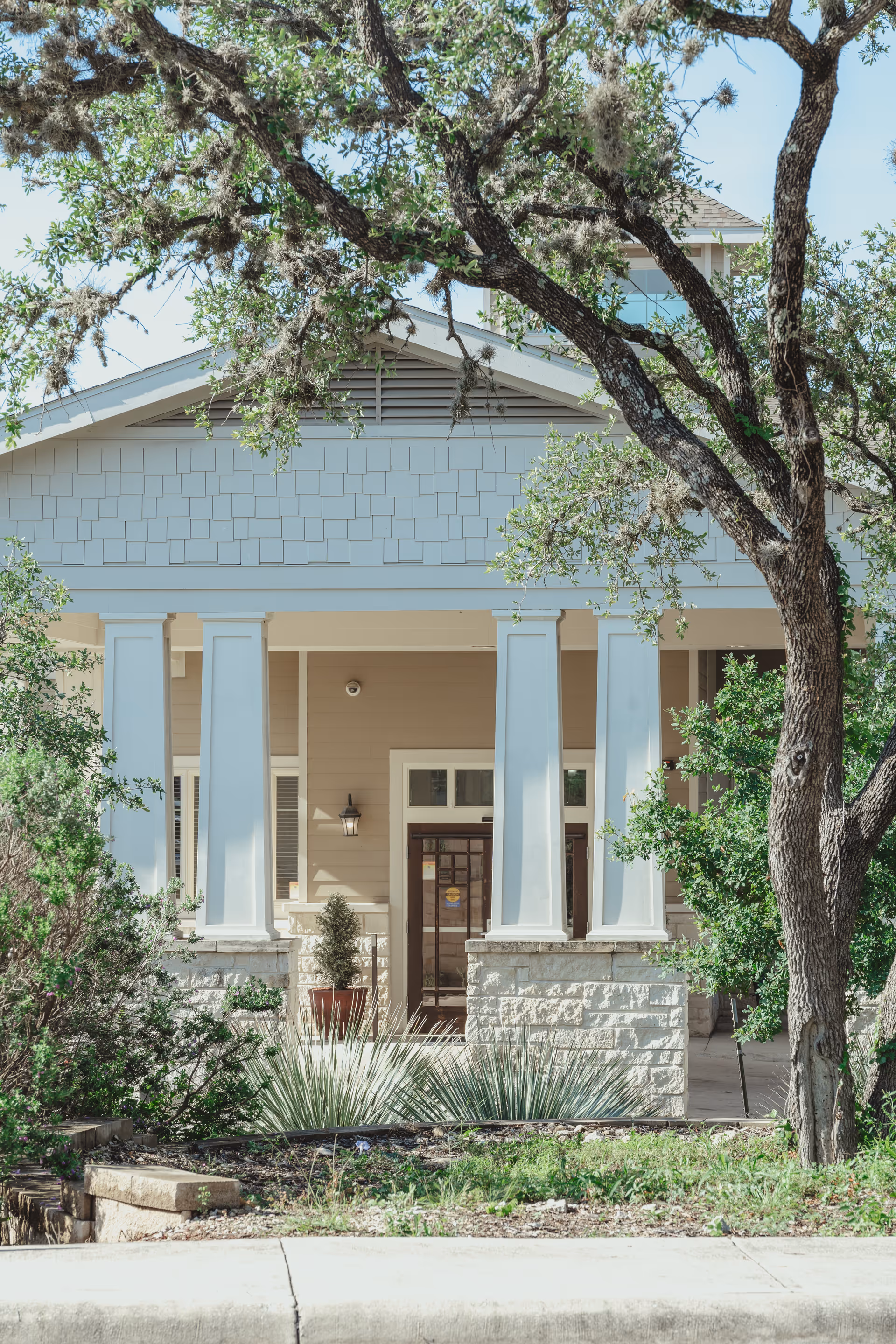 Front exterior view of a building with white pillars, beige siding, and stone accents, surrounded by trees and shrubs under a clear blue sky.