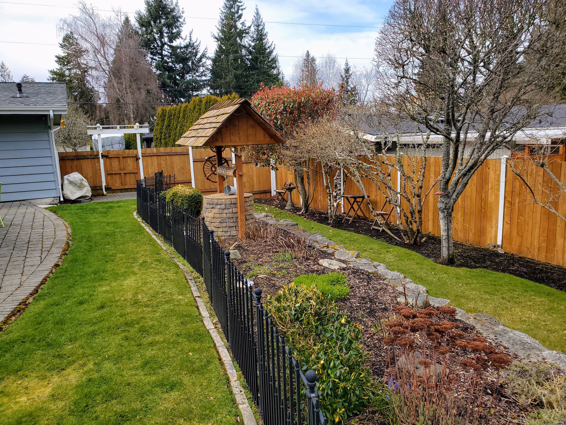 A well-maintained outdoor garden area with a green lawn, a black metal fence, various shrubs and plants, a wooden wishing well structure, and a wooden fence in the background. There are leafless trees and a small table with two chairs near the fence.