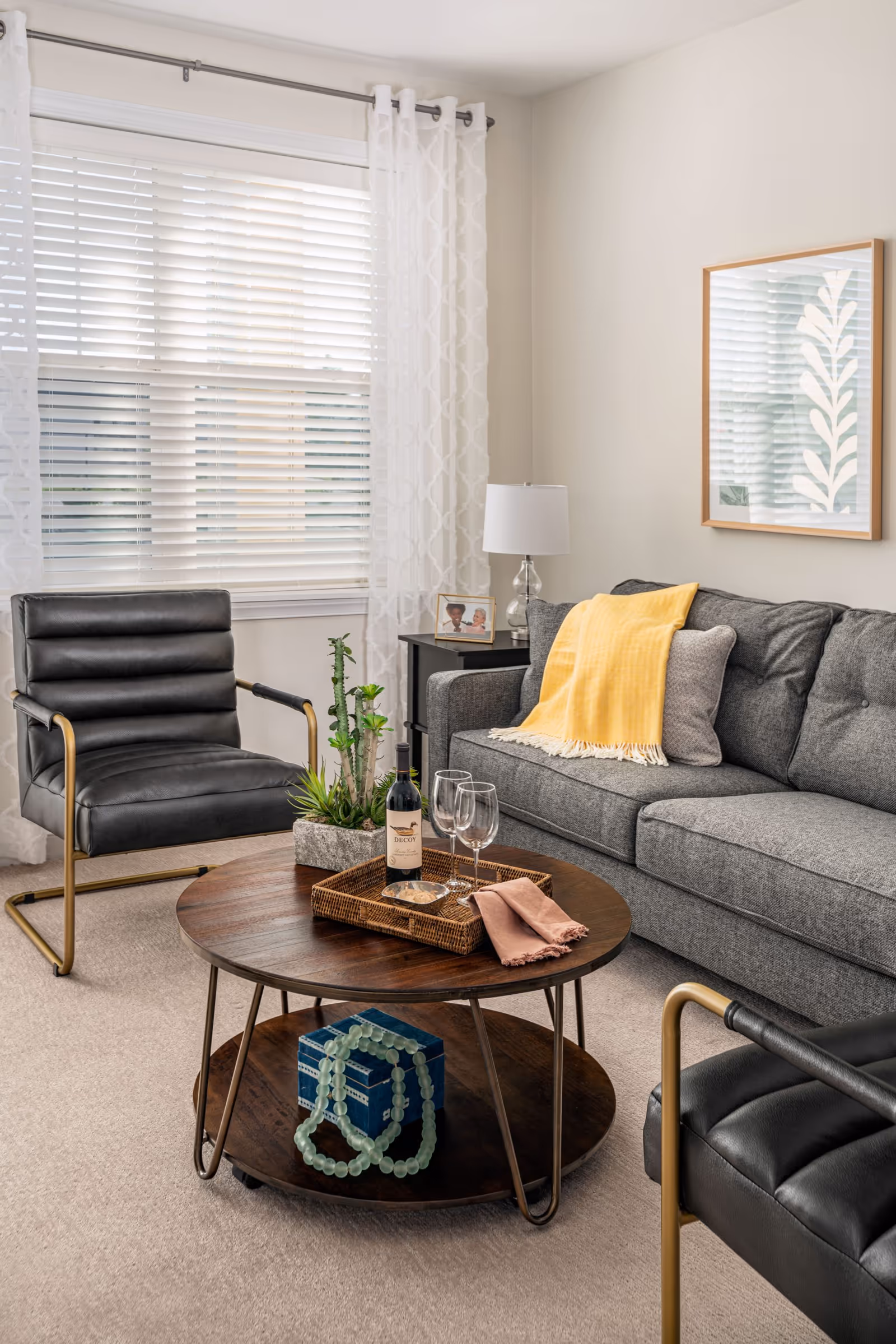A bright living room with a gray sofa draped with a yellow throw, black armchairs, and a round wooden coffee table set with wine and glasses in front of a large window with white blinds.