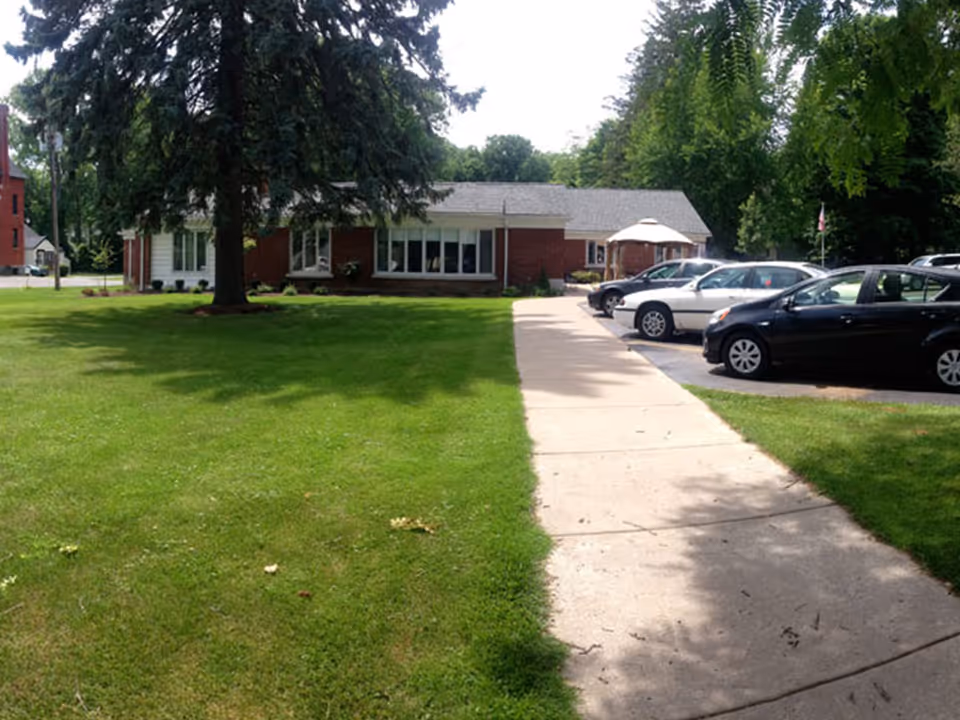 A single-story brick building with large windows, surrounded by a well-maintained green lawn and tall trees. A concrete sidewalk leads from the foreground to the building entrance. Several cars are parked in a parking area to the right of the sidewalk.