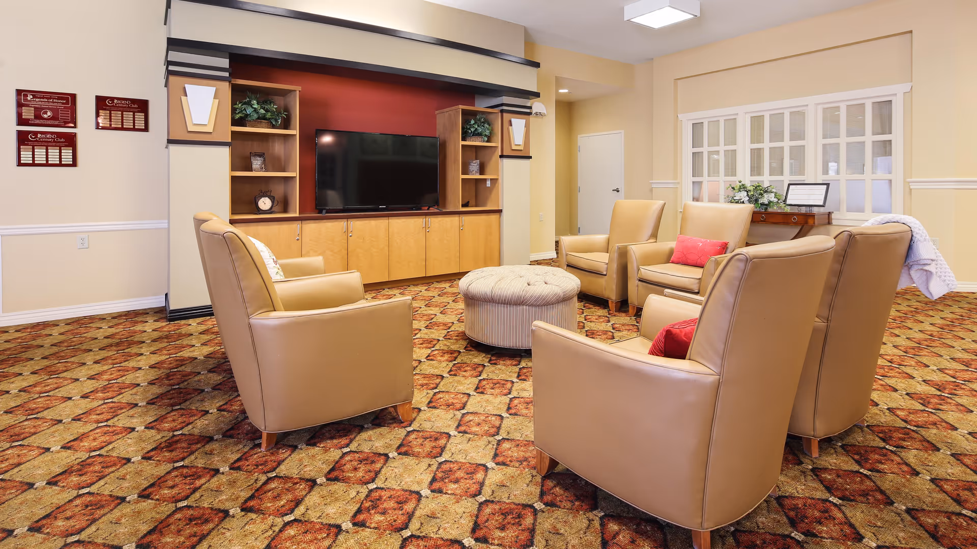 A cozy living room area with five tan leather armchairs arranged in a circle around a round, tufted ottoman. The room features a patterned carpet, a built-in wooden entertainment center with a flat-screen TV, and decorative plants on shelves. The walls are painted beige with white trim, and there is a window with white blinds and a small wooden table beneath it.