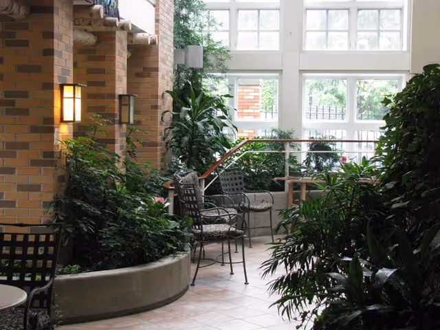 Indoor seating area with metal chairs and cushions surrounded by lush green plants and large windows letting in natural light. The space features brick walls with mounted lights and a tiled floor.