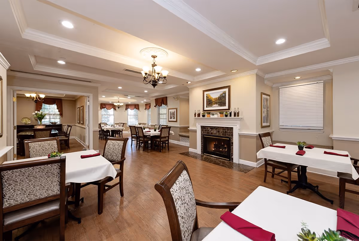A spacious dining room with multiple tables covered in white tablecloths and burgundy napkins. The room features wooden chairs with patterned upholstery, a fireplace with decorative items on the mantel, framed artwork on the walls, and large windows with valances allowing natural light to enter. The ceiling has recessed lighting and a chandelier.