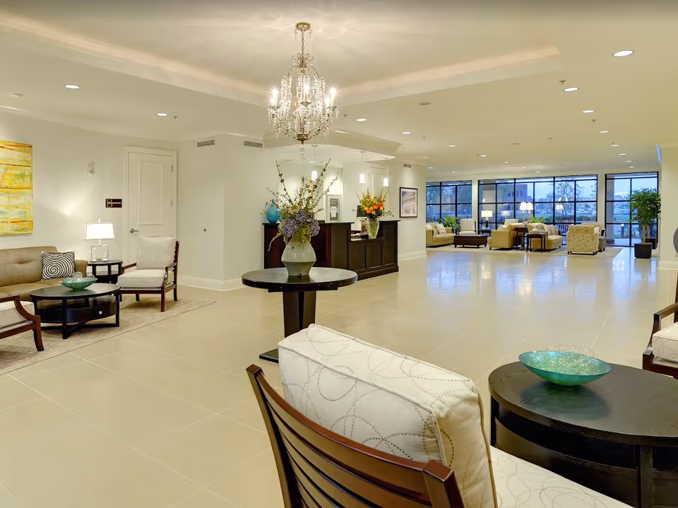 Spacious, well-lit senior living facility lobby with multiple seating areas, a central table with flowers, chandelier, and large windows.
