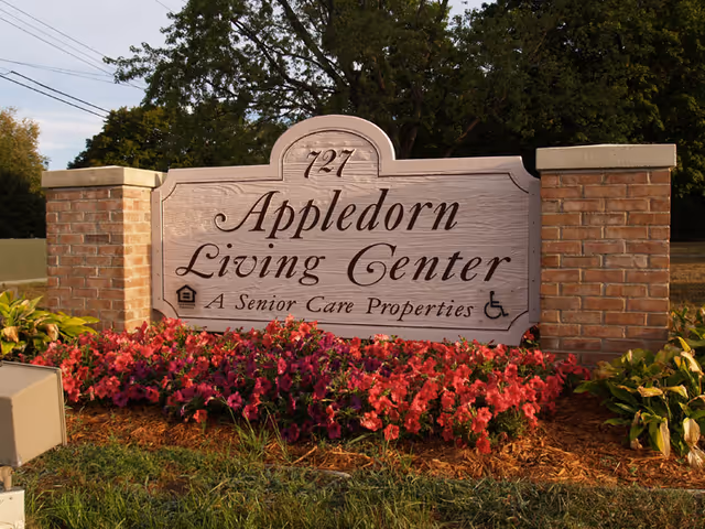 A large wooden sign with brick pillars on each side that reads '727 Appledorn Living Center A Senior Care Properties' surrounded by red flowers and greenery with trees in the background.