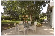 Patio area with a glass-top table and four chairs under a large tree beside a building.