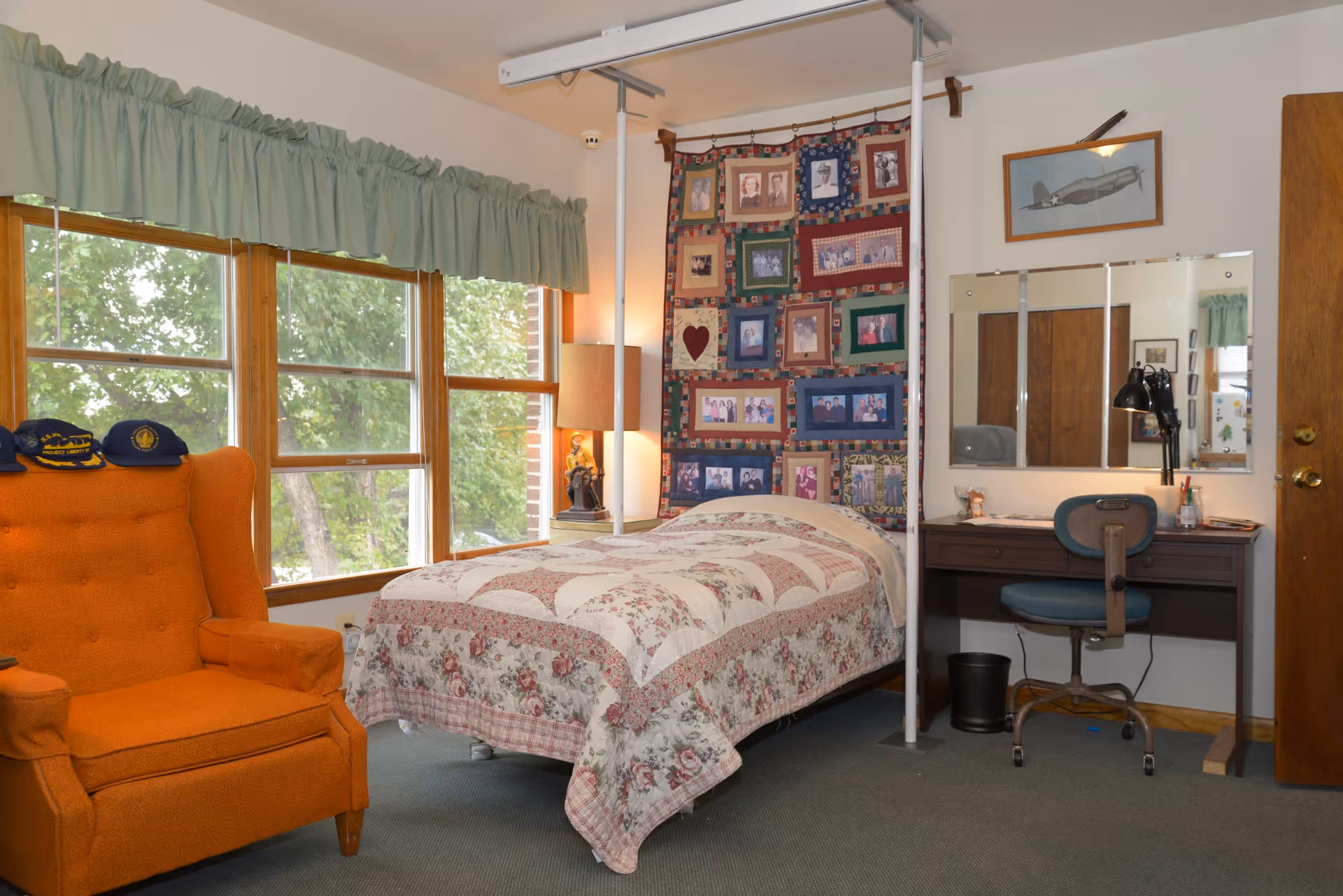 A cozy bedroom with a single bed covered in a floral and patchwork quilt. Behind the bed is a colorful quilt hanging on the wall featuring various framed photos. To the left of the bed is a large window with green curtains and an orange armchair with several hats on it. To the right of the bed is a wooden desk with a blue swivel chair, a desk lamp, and a large mirror above the desk. A framed picture of an airplane hangs on the wall above the mirror.