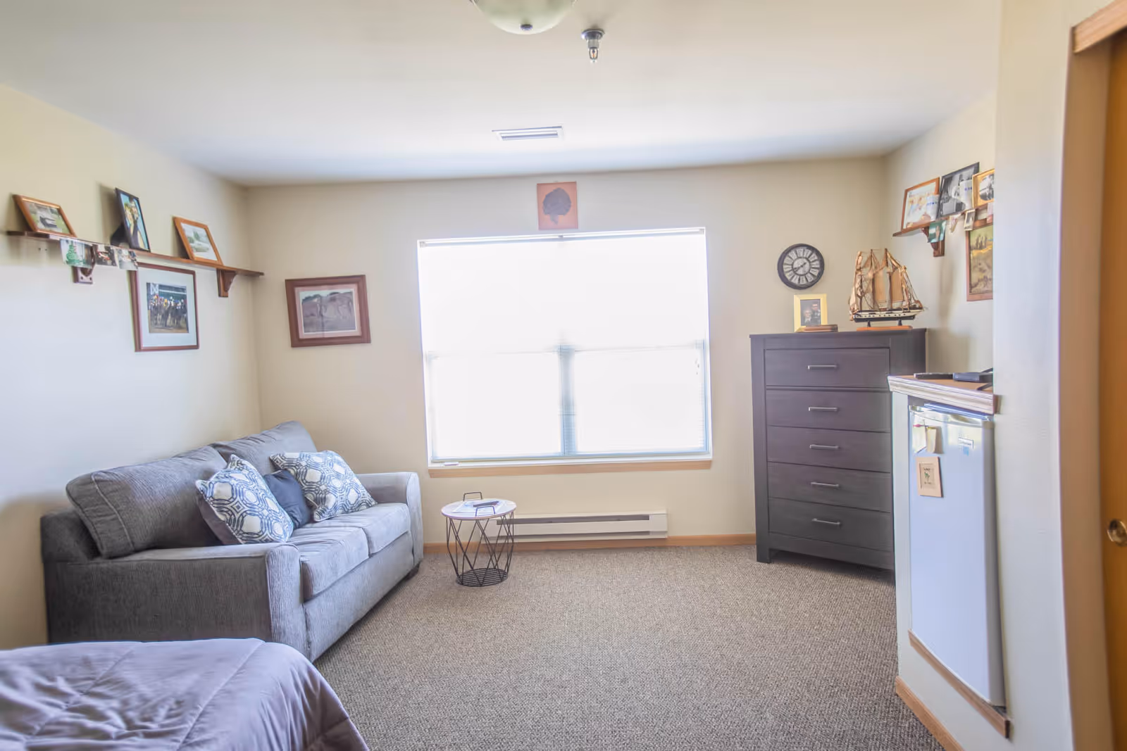 A bright and cozy living area in a senior living facility featuring a gray sofa with patterned cushions, a small round side table, a dark wooden chest of drawers with decorative items including a model ship and a clock, framed pictures on the walls, and a large window letting in natural light.