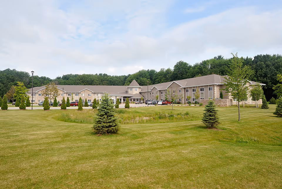 Exterior view of a large, two-story assisted living and memory care facility surrounded by a well-maintained lawn with small trees and shrubs, under a partly cloudy sky.