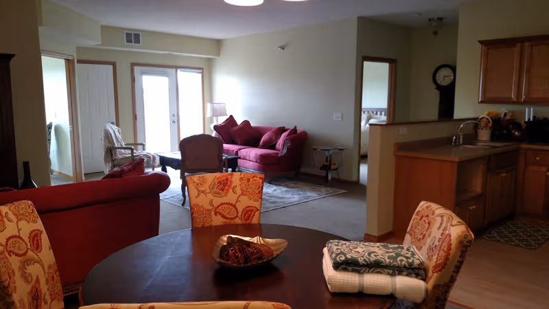 Open-plan living area with a dining table in the foreground, red sofas by a glass door and a kitchen to the right.