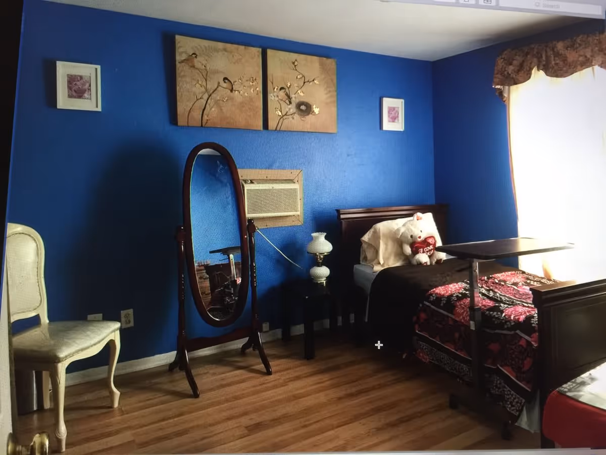 Bedroom with bright blue walls, a single bed topped with a teddy bear, an oval standing mirror, chair, bedside table and wood floor.