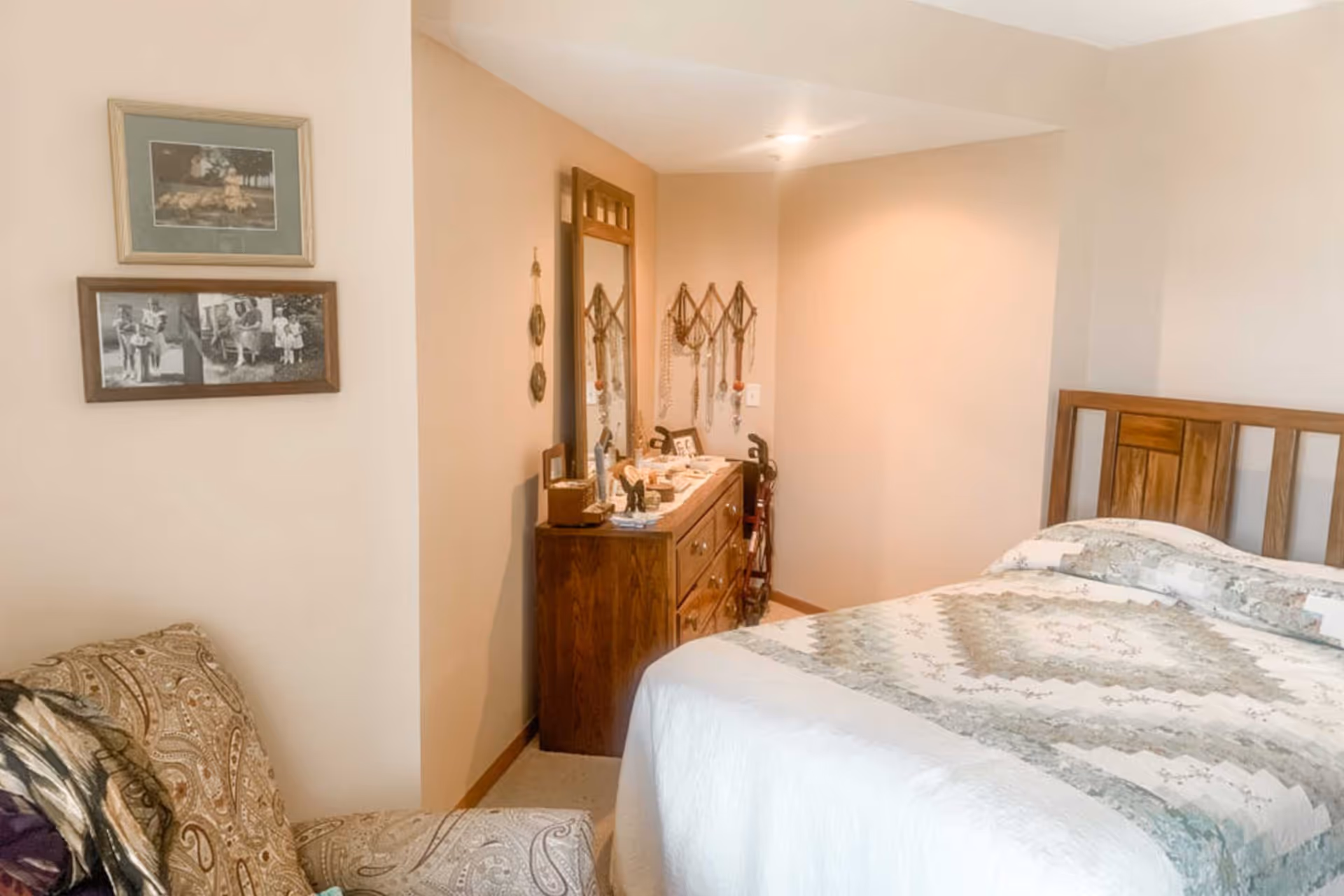 A cozy bedroom with a wooden bed frame and a quilted bedspread in soft colors. There is a wooden dresser with a mirror on top, decorated with various small items and jewelry hanging on the wall. A patterned armchair is partially visible in the foreground. The walls are painted a light beige color and two framed pictures hang on the wall.