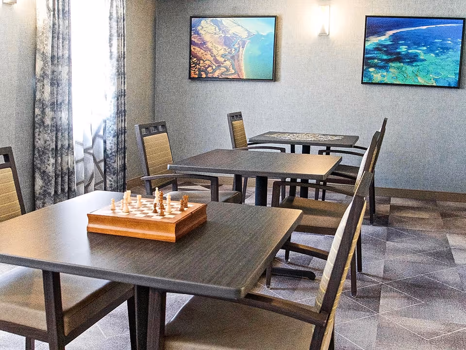 A room with several dark wooden tables and chairs arranged for seating. One table in the foreground has a chessboard with chess pieces set up. The room has patterned curtains on the window and two framed aerial landscape photographs on the wall.
