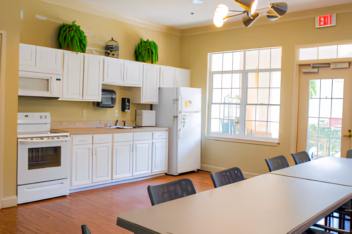 A bright kitchen area with white cabinets, a white stove, a white refrigerator, and a microwave. There are two green potted plants on top of the cabinets. In front of the kitchen is a long table with several black chairs. Large windows and a glass door allow natural light to fill the room.