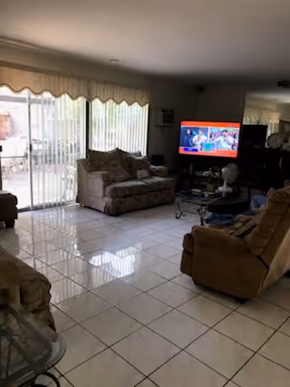 A living room with tiled floor, featuring a patterned sofa and a recliner chair. A flat-screen TV is on, placed on a stand against the wall. Large sliding glass doors with vertical blinds lead to an outdoor patio area. The room is softly lit with natural light coming through the glass doors.