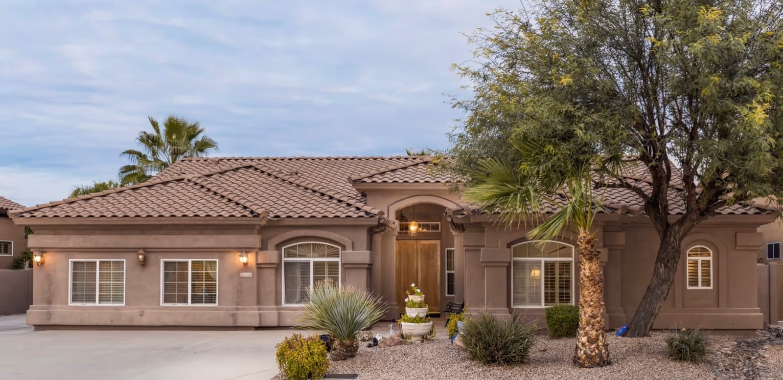 Front exterior view of a single-story building with a tiled roof, beige stucco walls, and several windows with white shutters. The entrance features a wooden door with a hanging light above it. The front yard has desert landscaping with gravel, various shrubs, a palm tree, and a larger tree on the right side.