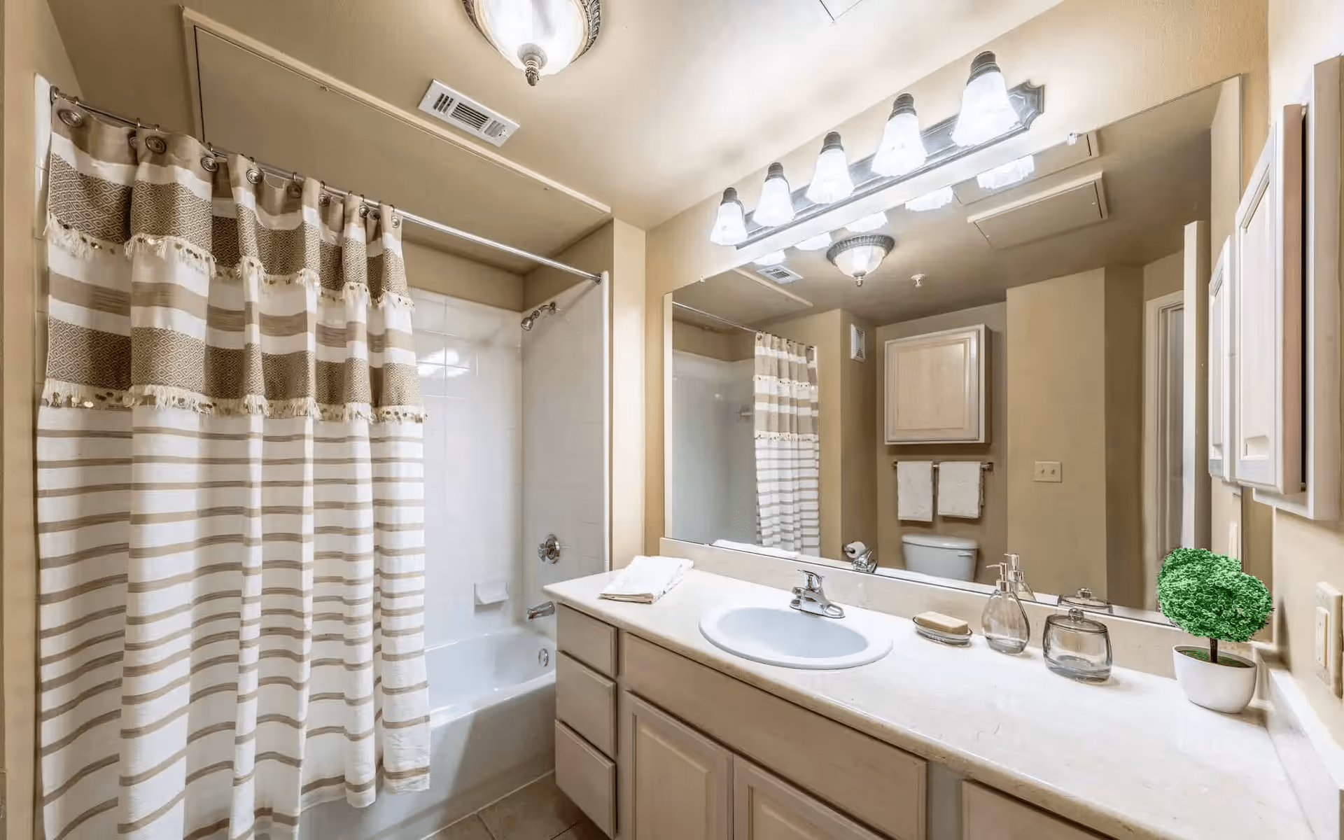 A bathroom with a white bathtub and shower combination with a beige and white striped shower curtain. There is a large mirror above a white countertop with a sink, soap dispenser, and a small potted plant. The walls are beige, and there are two towel racks with white towels and a wooden cabinet above the toilet.