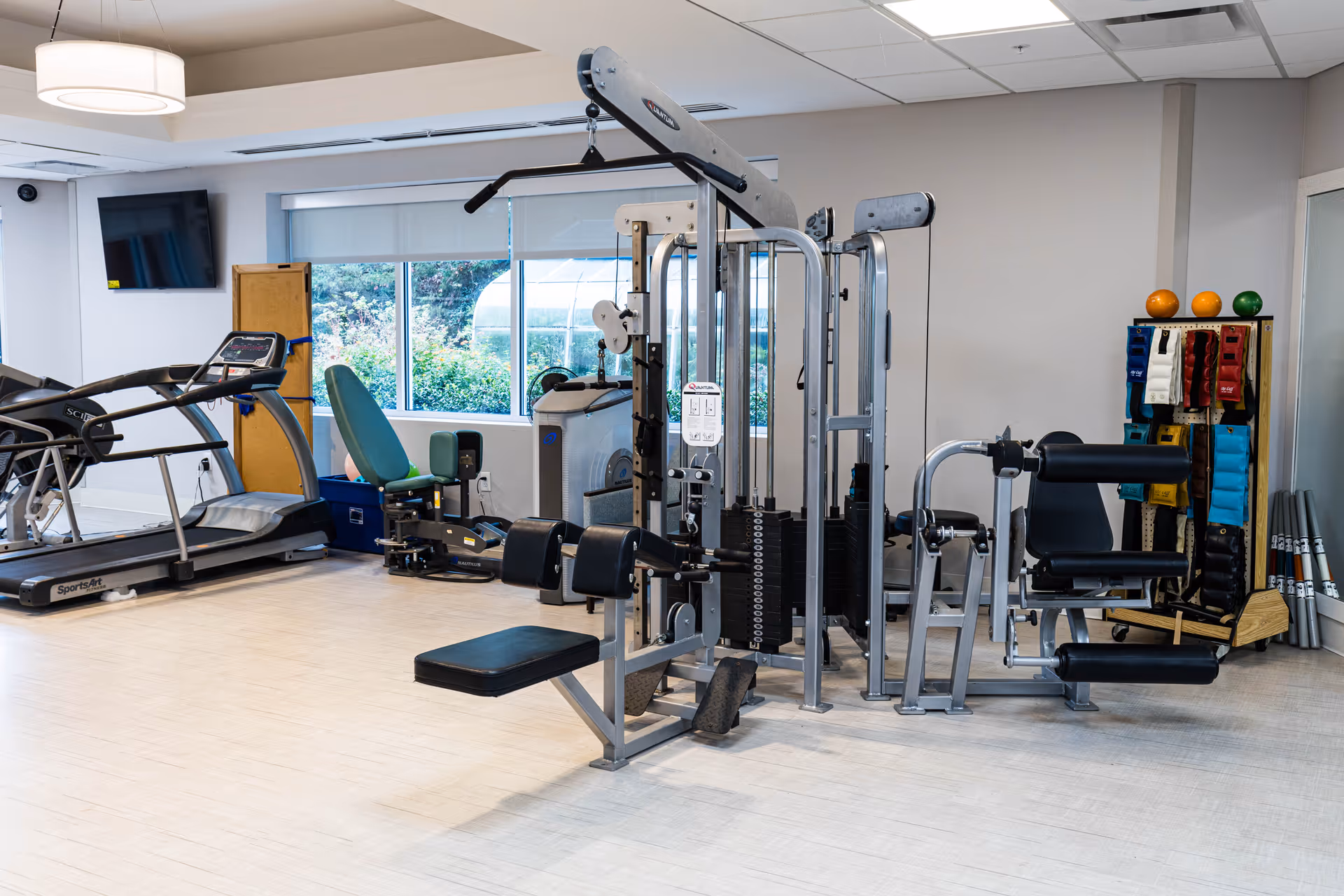 A fitness room with various exercise equipment including a treadmill, weight machines, and resistance bands. There is a large window showing greenery outside, a wall-mounted TV, and a light fixture on the ceiling.