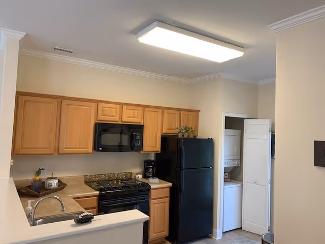 A compact kitchen area with light wood cabinets, a black microwave above a black stove, and a black refrigerator. There is a countertop with a sink and a few decorative items. To the right, there is a small laundry closet with a stacked washer and dryer behind white bi-fold doors. The walls are painted beige and the ceiling has a rectangular fluorescent light fixture.