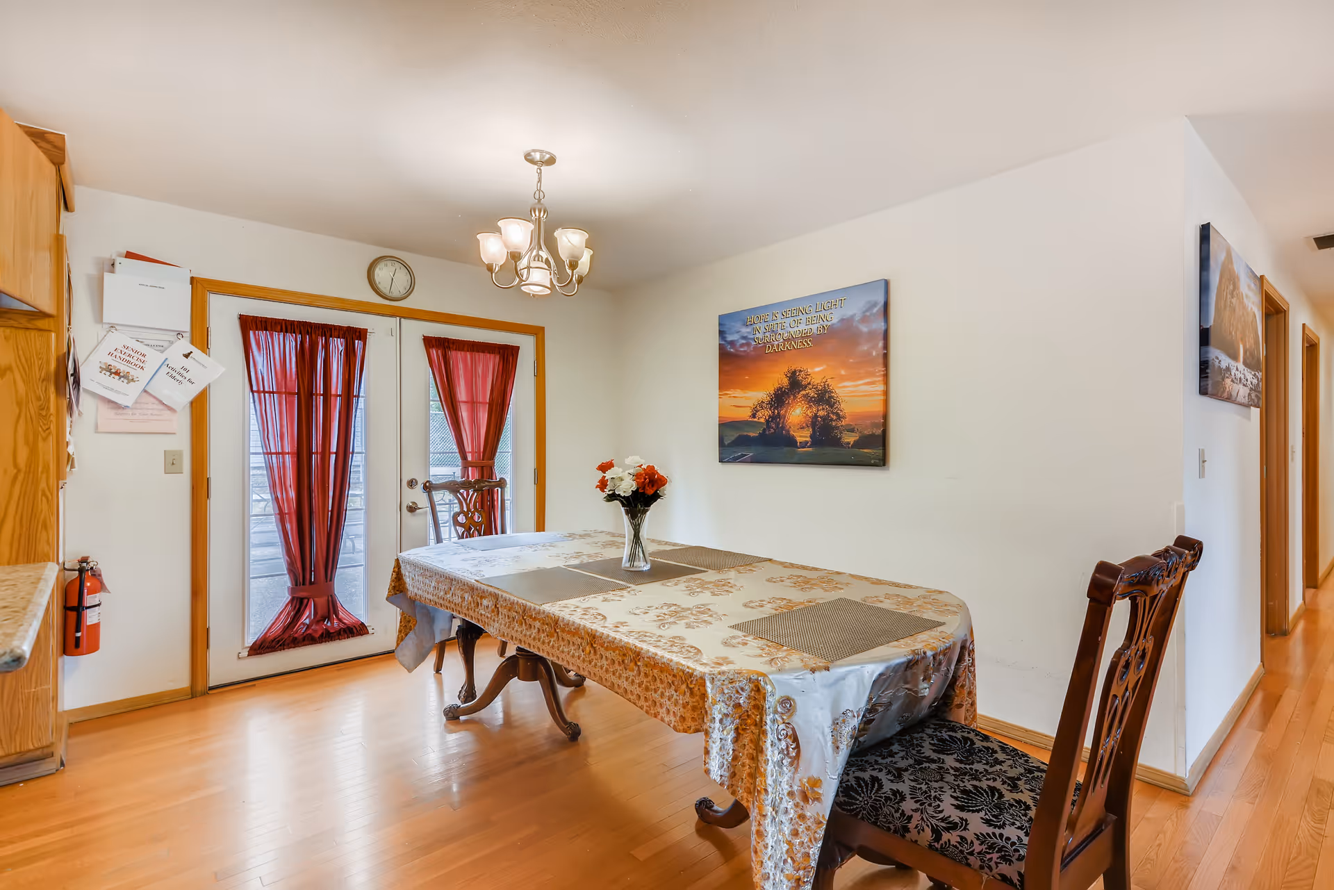 A dining room with a wooden table covered by a floral tablecloth and a vase with flowers in the center. There are two wooden chairs with patterned cushions around the table. Behind the table, there are double glass doors with red curtains, a clock above the doors, and a wall with a framed picture showing a sunset and an inspirational quote. The floor is wooden, and a chandelier with five lights hangs from the ceiling.