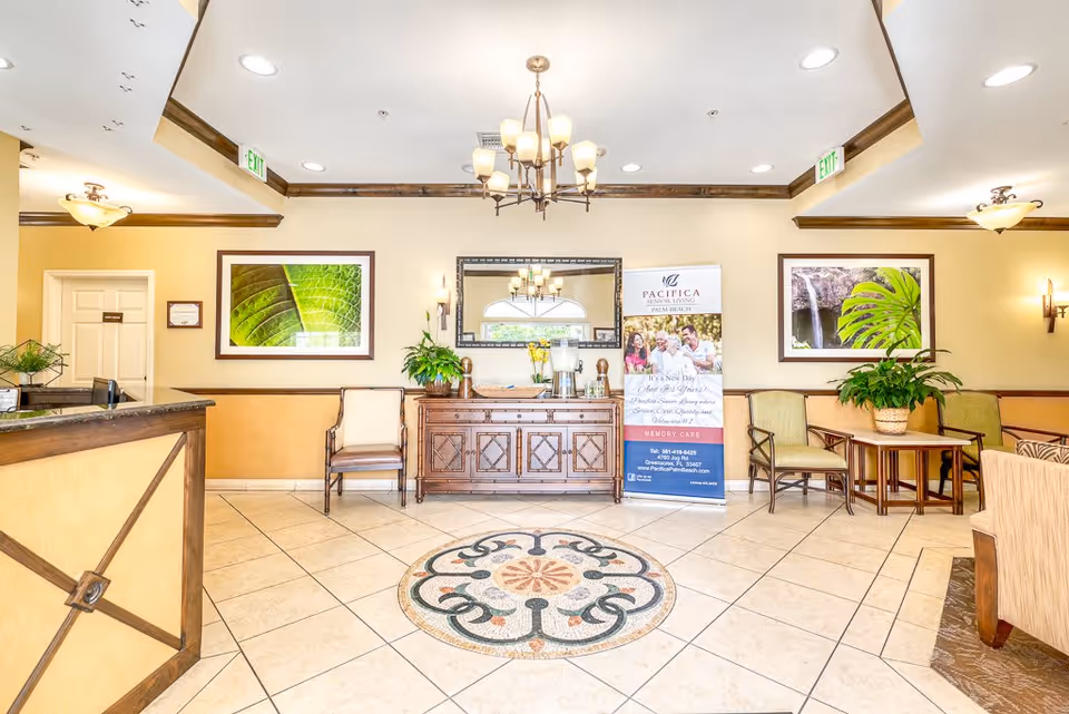 A bright and welcoming senior living facility lobby with tiled floors featuring a decorative circular pattern in the center. The walls are painted in warm tones with dark wood trim. There is a wooden reception desk on the left, a wooden sideboard with plants and a coffee station in the center, and a seating area with chairs and a small table on the right. Two large framed nature photographs hang on the walls, and a chandelier and ceiling lights illuminate the space. A banner for Pacifica Senior Living Palm Beach is displayed near the sideboard.