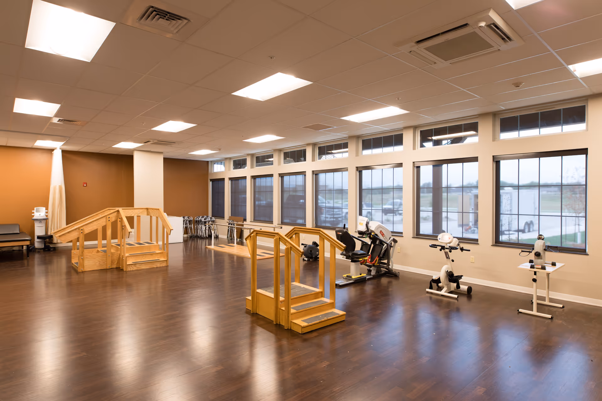 A spacious rehabilitation or physical therapy room with wooden handrails and steps for mobility exercises, exercise bikes, and large windows letting in natural light.