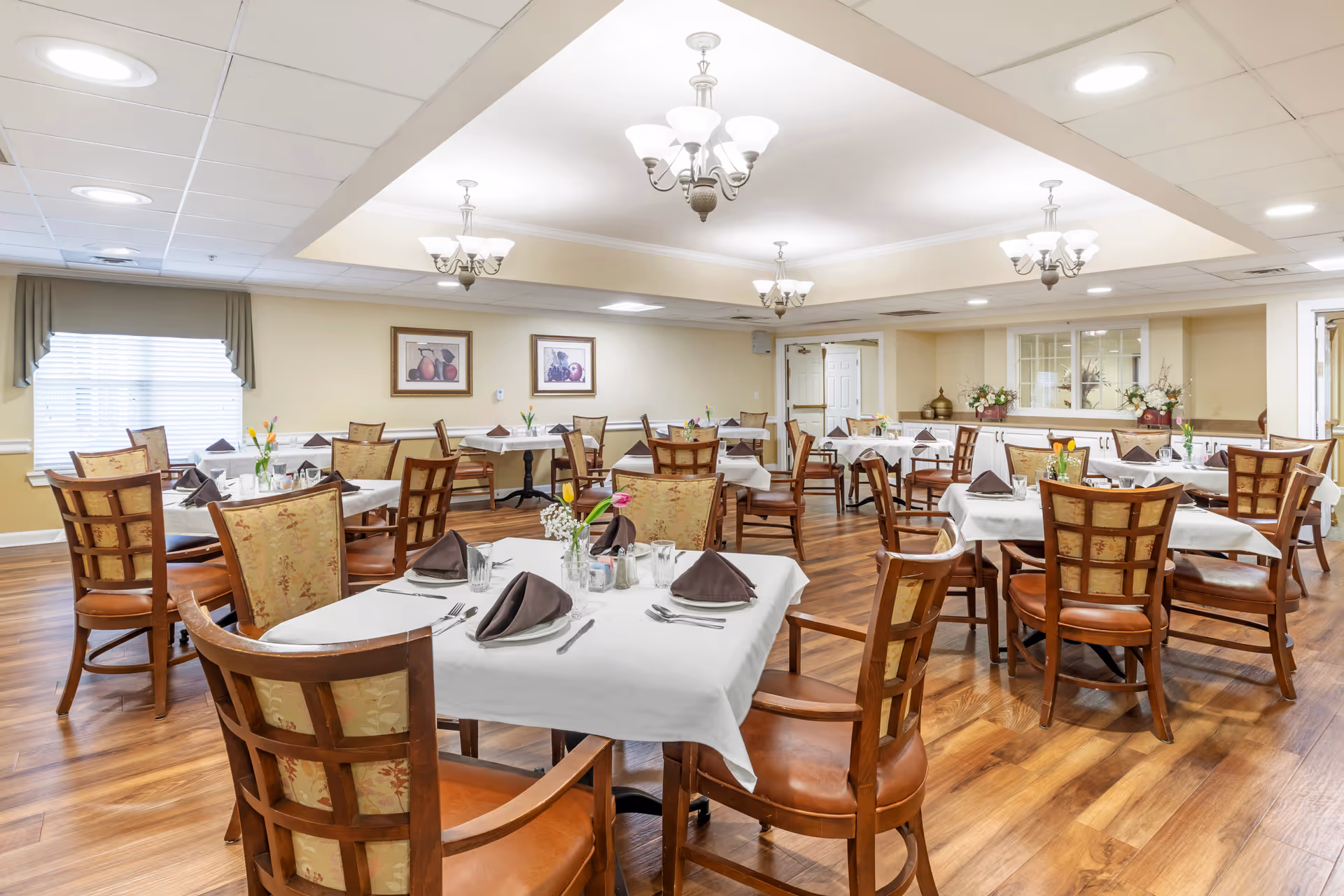 A well-lit dining room with multiple tables covered in white tablecloths, each set with folded brown napkins, glasses, and silverware. The room features wooden chairs with cushioned seats and backs, hardwood flooring, chandeliers hanging from the ceiling, and framed artwork on the walls. There are windows with curtains and floral centerpieces on the tables.