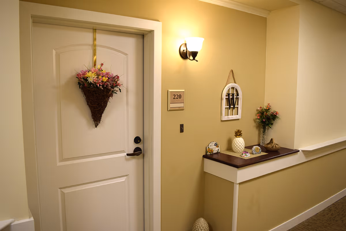 Hallway view showing a decorated apartment door numbered 220 with a wall sconce and a small shelf holding decorative items.