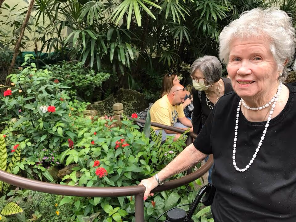 An elderly woman with white hair wearing a black top and a white beaded necklace is smiling and holding onto a railing in a lush indoor garden. Behind her, several other people, including an elderly woman wearing a face mask, are observing the plants and flowers. The garden is filled with green foliage and red flowers.