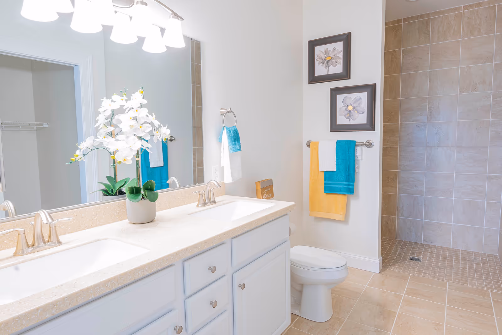 Bright bathroom with a double-sink vanity and large mirror, an orchid on the counter, a toilet, colorful towels, and a tiled walk-in shower.
