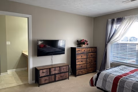 A bedroom with a bed covered in a striped comforter in red, gray, and white. There is a window with sheer and dark curtains on the right side, allowing natural light to enter. Against the wall, there is a flat-screen TV mounted above a wooden dresser with multiple drawers. Next to it, there is a taller wooden chest of drawers with a flower arrangement on top. An open doorway leads to another room with a tiled floor.