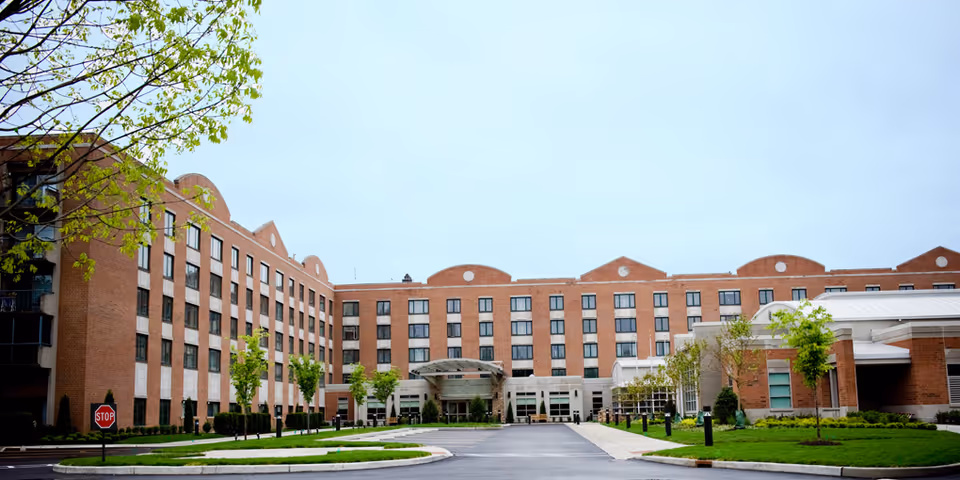 Front exterior view of a multi-story red-brick senior living facility with an entrance canopy, driveway, and landscaped grounds.