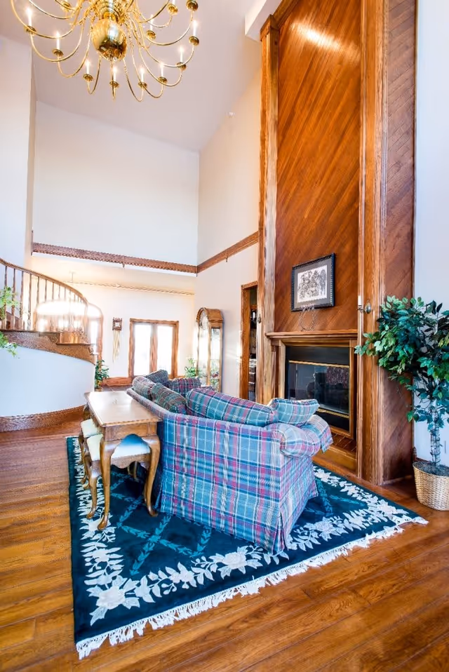 A cozy living room with a tall wooden fireplace, a plaid sofa, a wooden side table with stools underneath, a blue floral rug, a large chandelier, and a potted plant. There is a staircase with wooden railing and a large window letting in natural light.