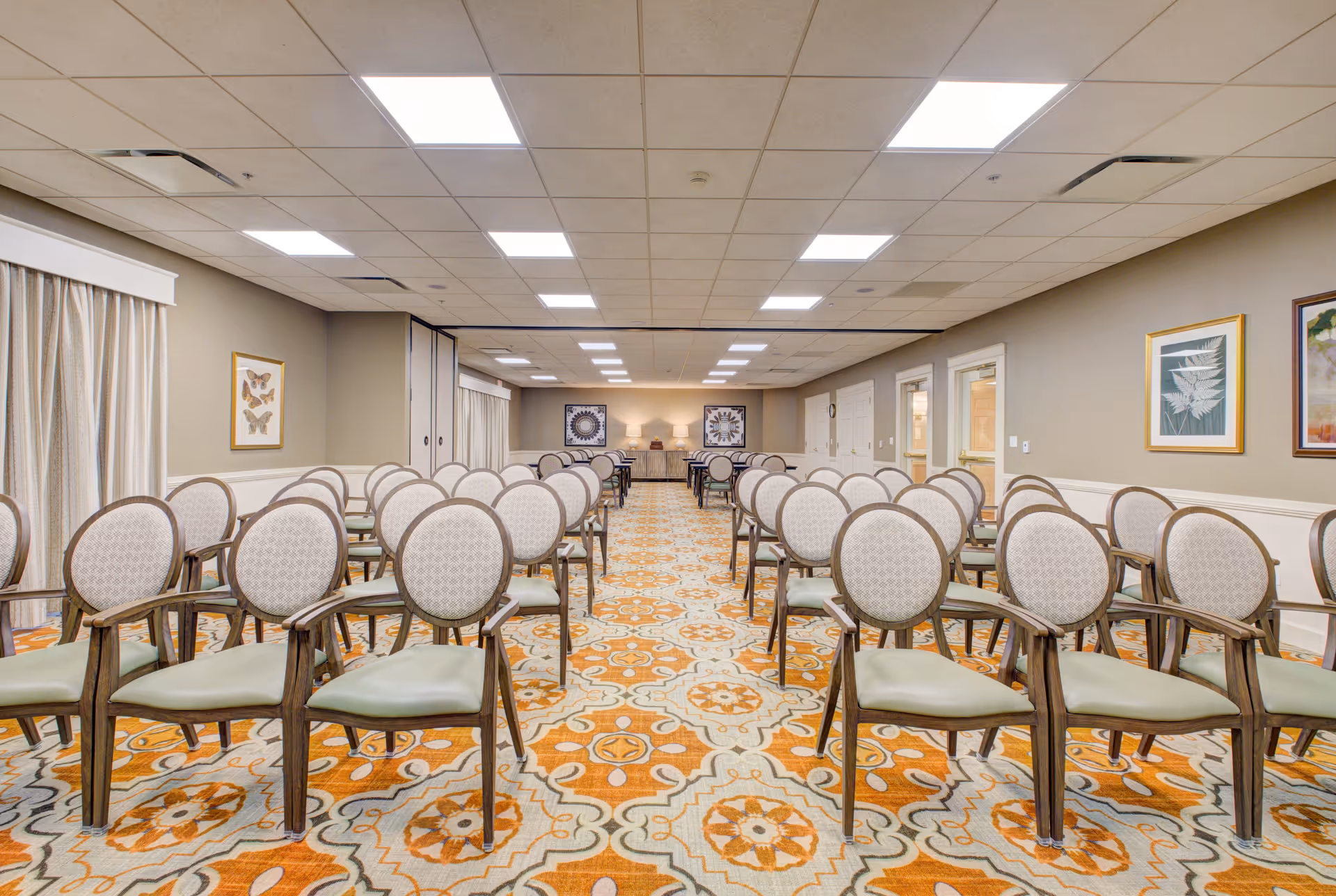 A large room set up with multiple rows of chairs arranged in pairs facing forward. The room has a patterned carpet with orange, beige, and white colors, beige walls adorned with framed artwork, and a ceiling with recessed lighting panels. There are curtains along one side and doors along the other side of the room.