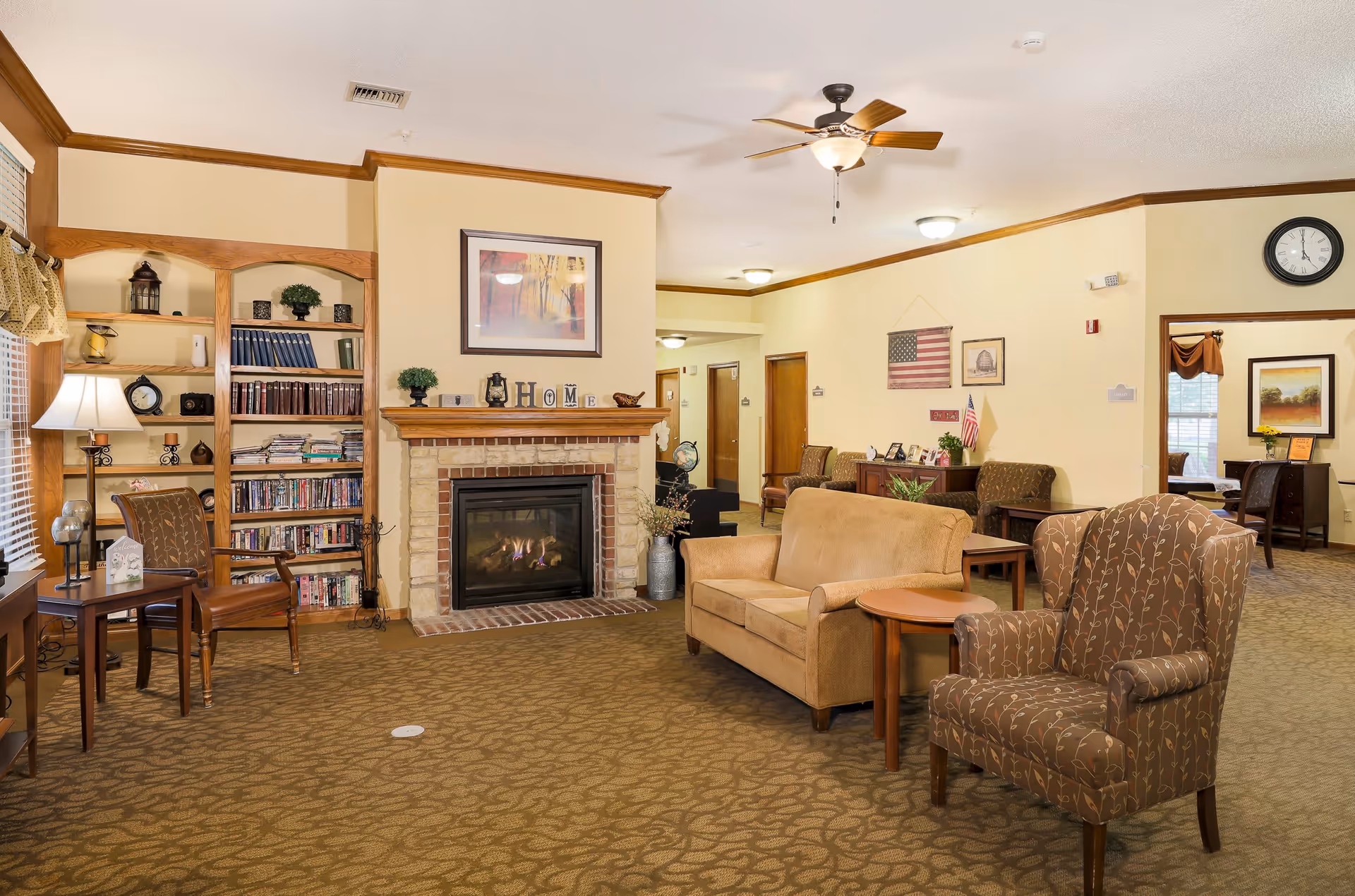 A cozy living room area in an assisted living facility featuring a fireplace with a 'HOME' decoration on the mantle, a bookshelf filled with books and decorative items, a beige loveseat, a patterned armchair, side tables, and a ceiling fan. The room has warm lighting, carpeted floor, and walls adorned with framed pictures and an American flag.