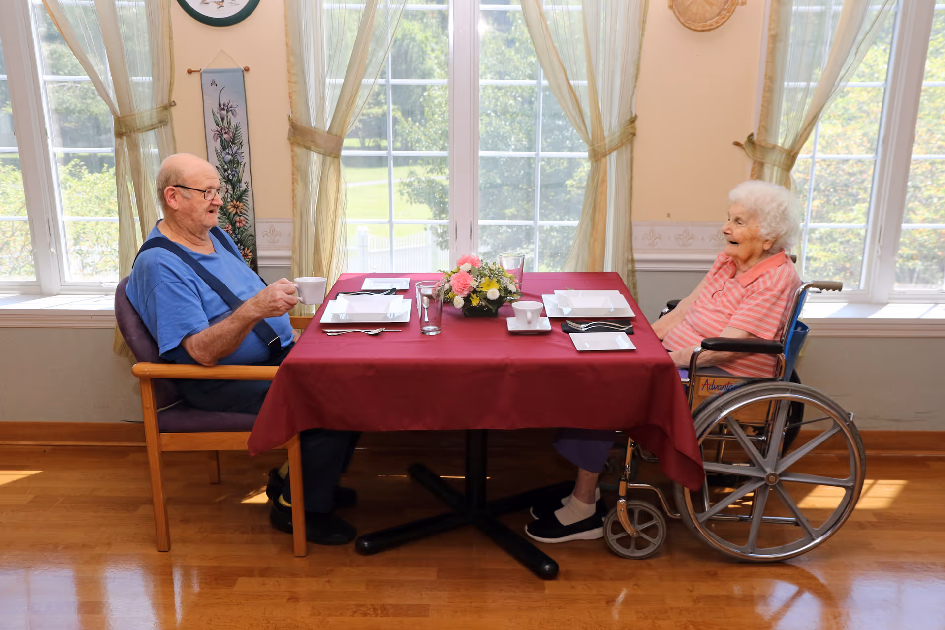 An elderly man and woman sitting across from each other at a dining table with a red tablecloth in a room with large windows and sheer curtains. The man is holding a cup, and the woman is seated in a wheelchair. The table is set with plates, cups, and a floral centerpiece.