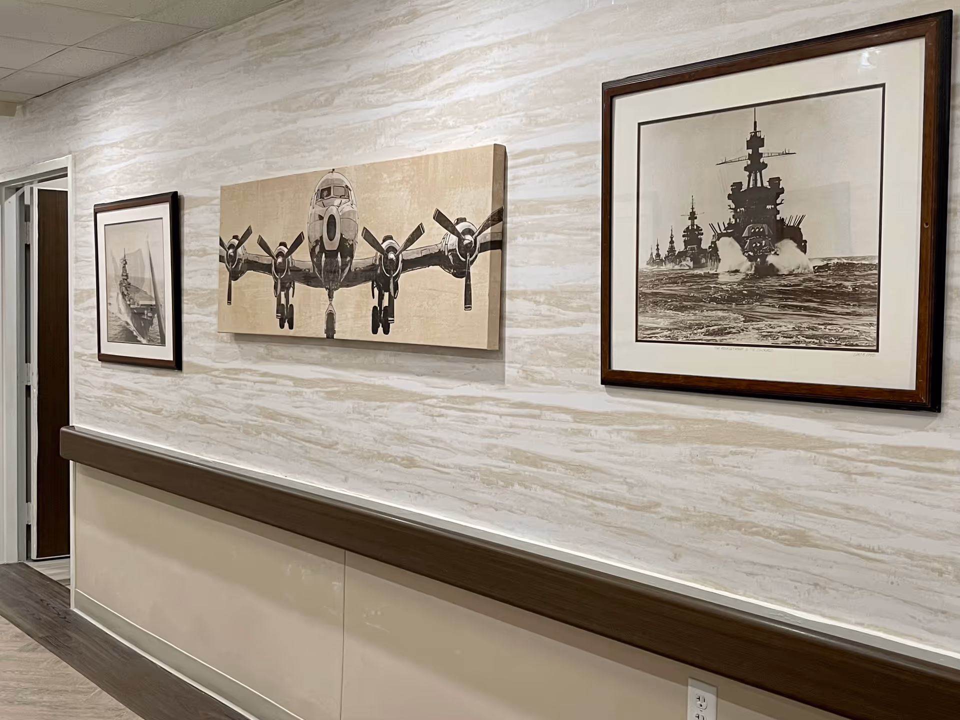 A hallway with light-colored textured walls featuring three framed artworks: a front view of a propeller airplane in the center and two images of battleships on either side. The hallway has a wooden handrail and a door at the end.