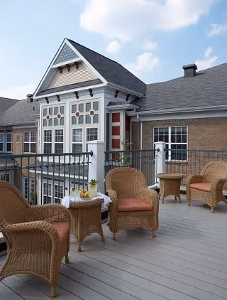 Outdoor patio area with wicker chairs and small tables on a wooden deck, attached to a brick building with large windows and a decorative gable roof under a partly cloudy sky.