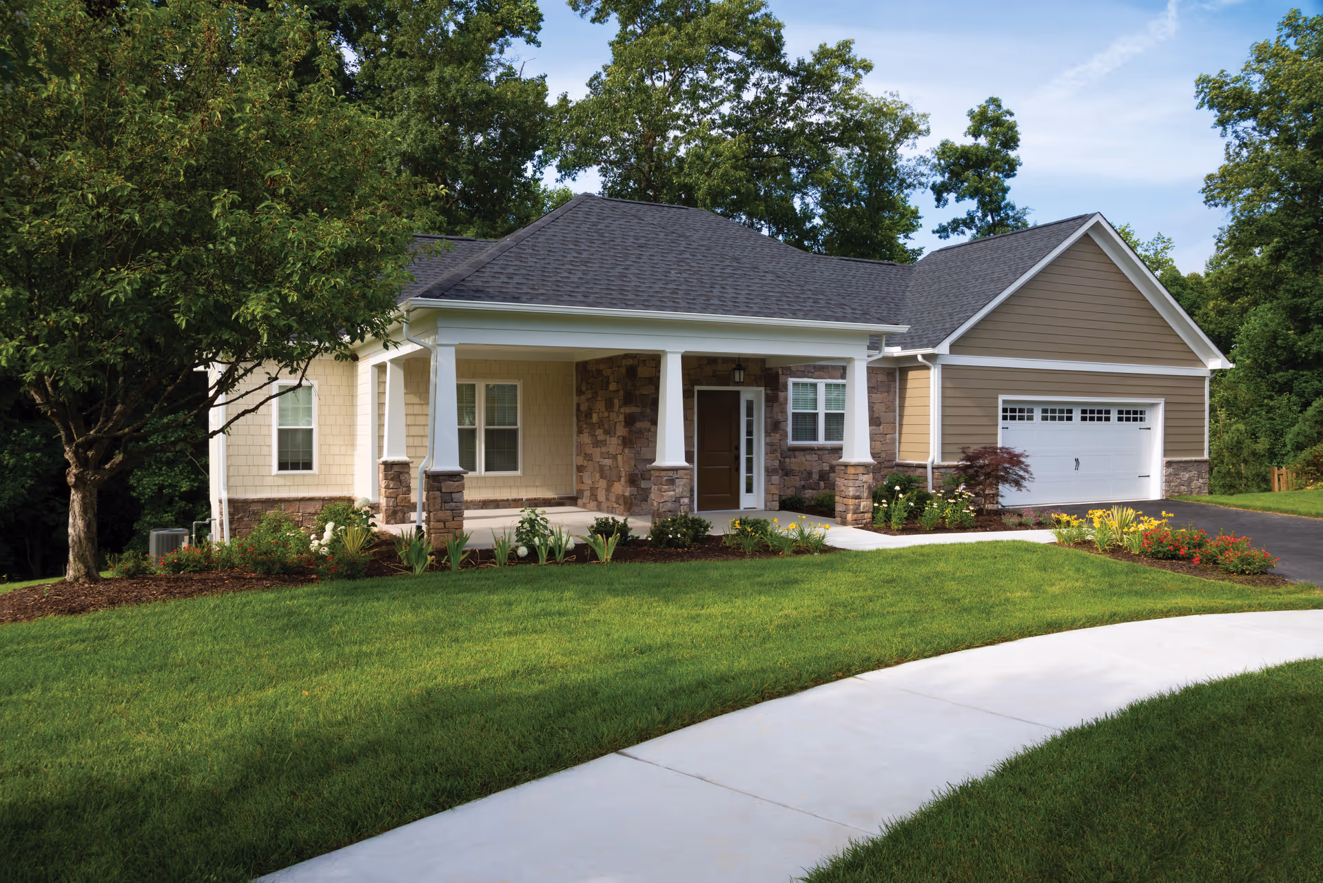 A single-story house with a covered front porch supported by white columns with stone bases. The house has beige and brown siding, a dark shingled roof, and a white garage door. There is a well-maintained lawn, a curved concrete walkway leading to the porch, and various shrubs and flowers planted around the house. Trees are visible in the background under a partly cloudy sky.