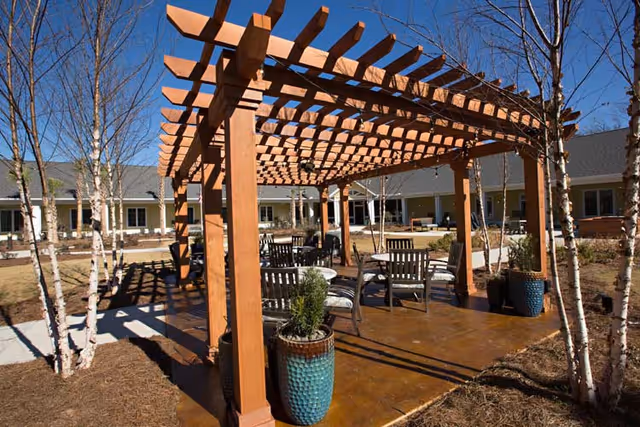 Outdoor seating area with wooden pergola, several chairs and tables arranged underneath, surrounded by small trees and plants in large pots, with a building visible in the background under a clear blue sky.