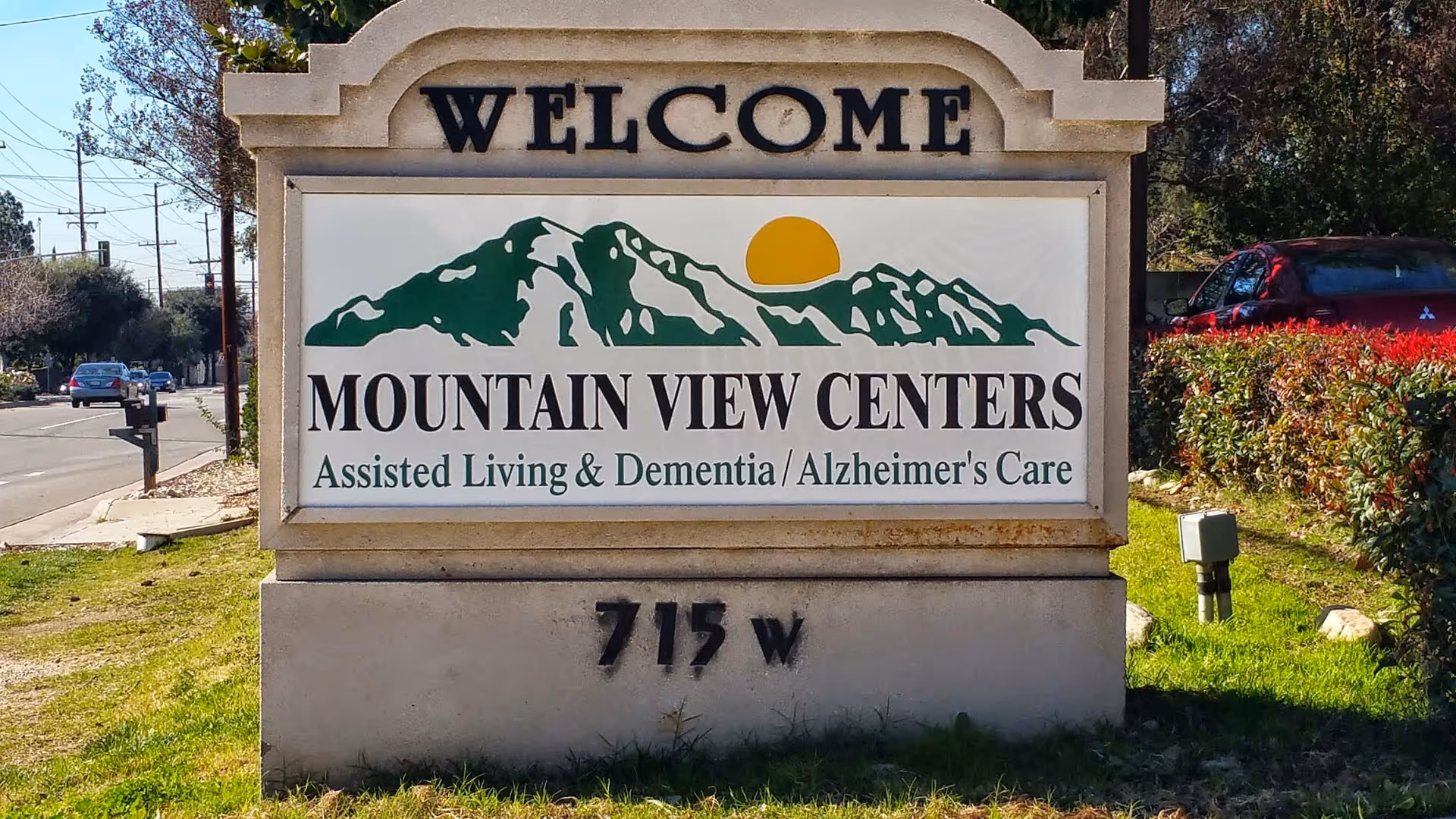 Outdoor stone sign with the text 'WELCOME' at the top, followed by a graphic of green mountains with a yellow sun, and the words 'MOUNTAIN VIEW CENTERS Assisted Living & Dementia / Alzheimer's Care' below. The sign is located on a grassy area next to a street with cars and bushes visible in the background.