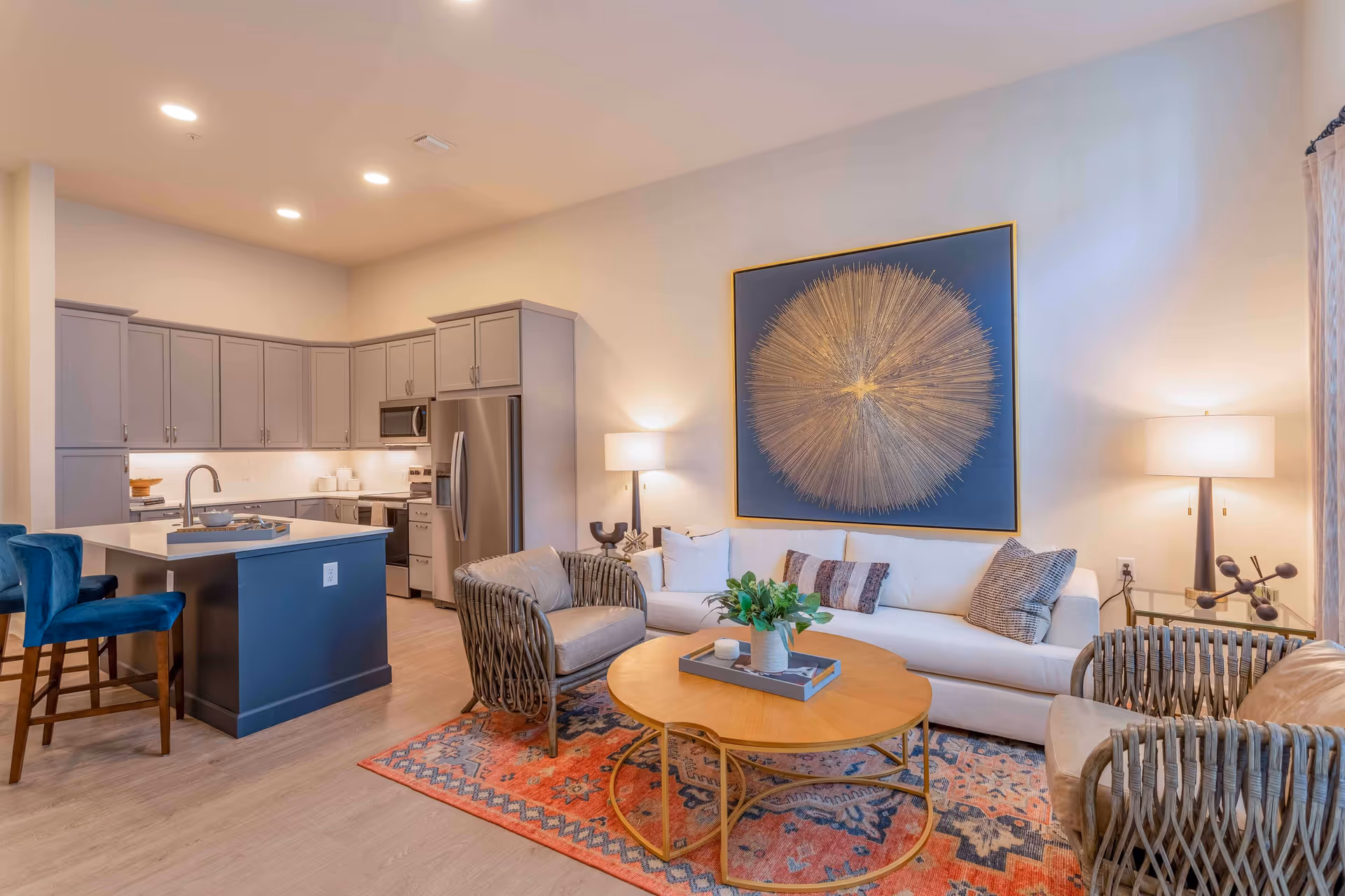 A modern open-concept living room and kitchen area featuring a white sofa with decorative pillows, two wicker armchairs with cushions, a round wooden coffee table with a plant centerpiece, and a colorful patterned rug. The kitchen has gray cabinets, stainless steel appliances, a blue island with a sink, and two blue bar stools. The walls are light-colored with a large abstract circular artwork above the sofa and two table lamps providing warm lighting.