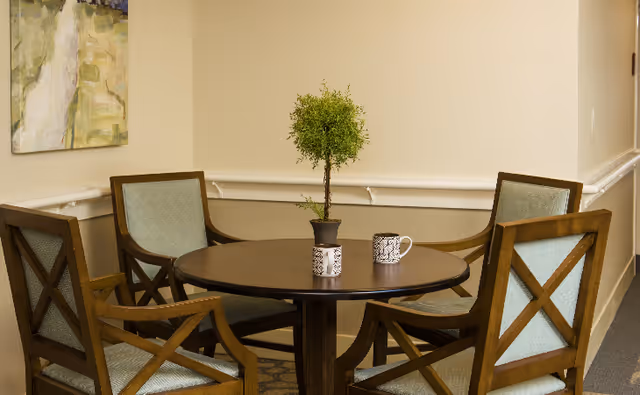 Round wooden table with a small potted plant and two mugs surrounded by four wooden chairs in a neutral seating area.