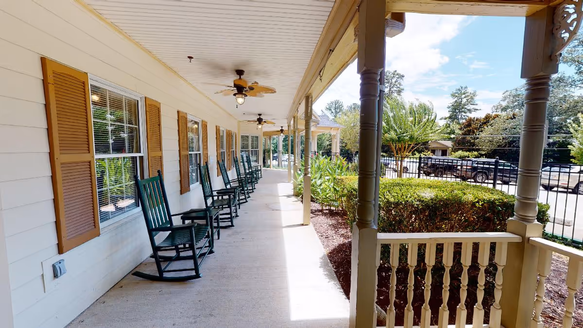 A covered outdoor porch area with multiple dark green rocking chairs lined up against a cream-colored wall with windows and brown shutters. The porch has ceiling fans and overlooks a garden area with bushes and trees, with a parking lot and cars visible in the background under a partly cloudy sky.