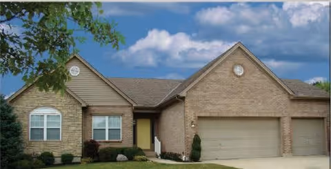 Single-story brick house with a three-car garage, a small front porch, and a well-maintained lawn under a blue sky with some clouds.