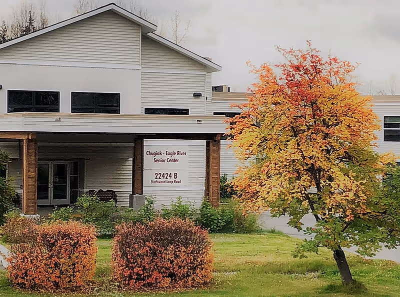 Exterior view of the Chugiak - Eagle River Senior Center building with a covered entrance supported by wooden posts. There is a sign displaying the facility name and address: 22424 B Birchwood Loop Road. In front of the building, there are bushes with autumn-colored leaves and a tree with yellow and orange foliage.